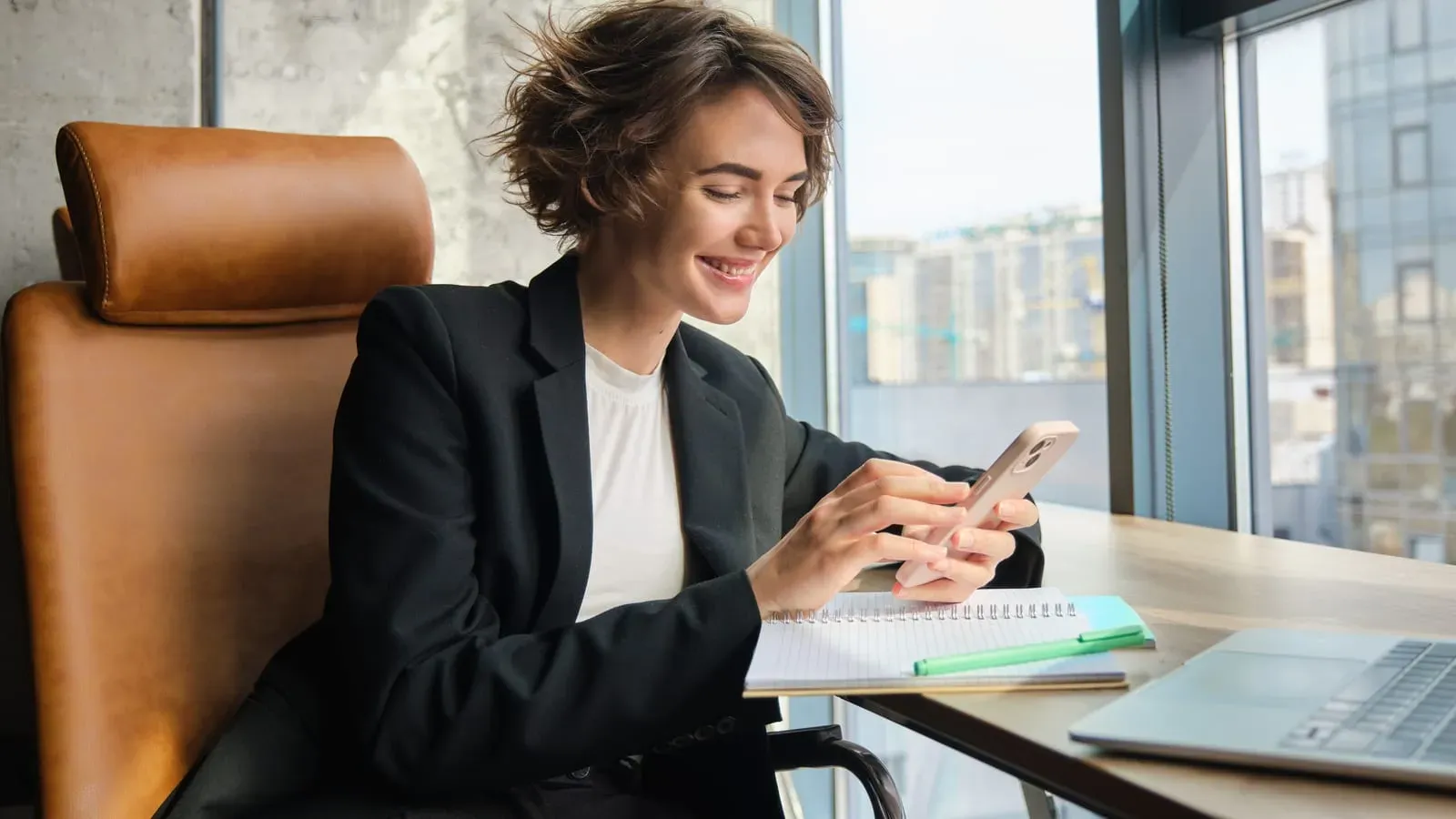 Profissional sorrindo enquanto responde mensagens no celular, sentada à mesa do escritório